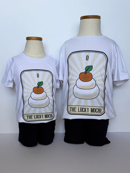 This is a photograph of a toddler mannequin and a youth mannequin.  Both are wearing white t-shirts with a central design.  The design is of a tarot card with the Roman numeral zero and the text "The Lucky Mochi" on the bottom.  The central image on the tarot card is of two white mochis stacked one on top of the other, with a tangerine on the top.  